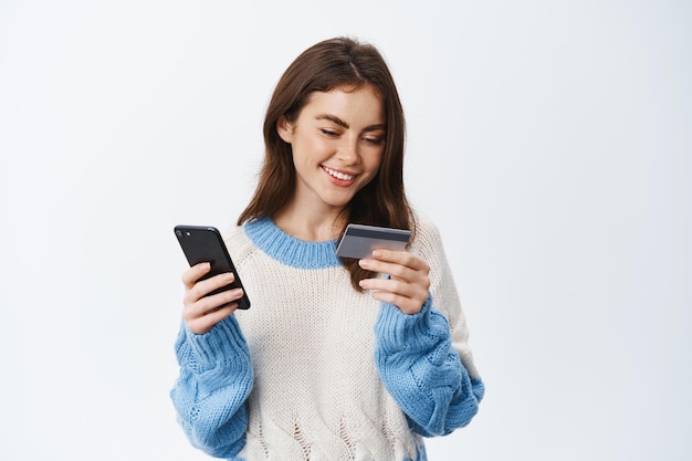 Photo young smiiling girl online shopping or sending money via mobile app, holding plastic credit card and smartphone, standing on white