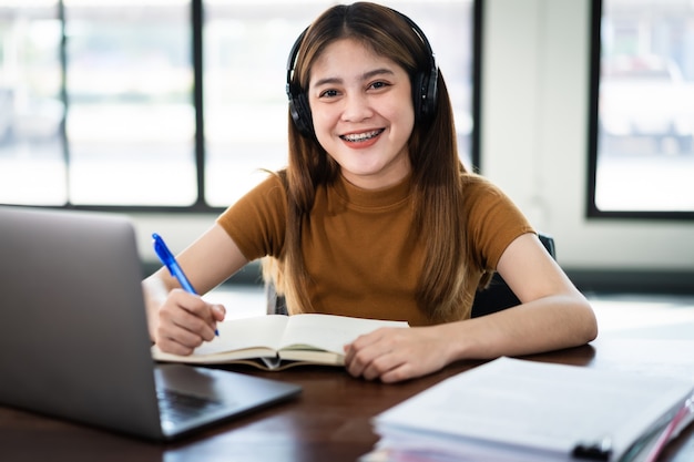 若い笑顔のアジアの女子学生は、ワイヤレスヘッドホンを着用してノートブックに書き込み、言語をオンラインで学習し、講師の話を聞きます。自宅でのビデオ通話のeラーニングによるウェビナー、遠隔教育