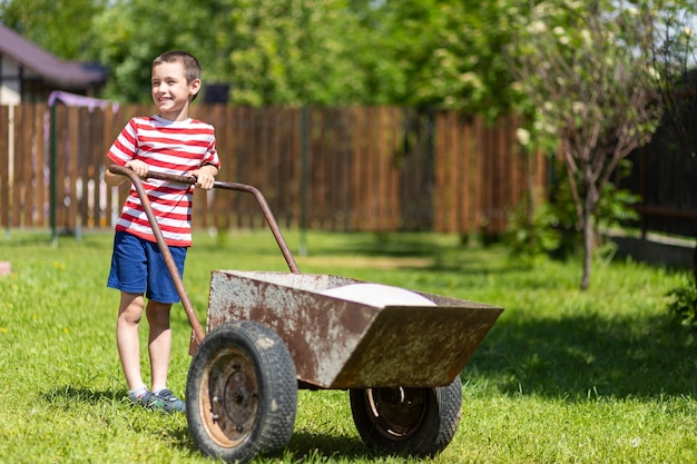 若い笑顔の男の子が庭の周りに手押し車を押します。Tショーツとショートパンツで楽しんでいる男の子のヘルパー