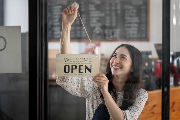 カフェの窓ガラスに掛かっているオープン看板を保持している若い笑顔の女性起業家