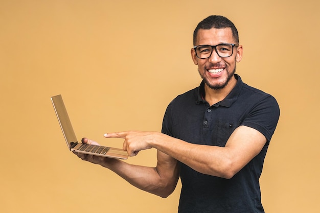 Giovane uomo afroamericano sorridente in piedi e utilizzando il computer portatile isolato su sfondo beige