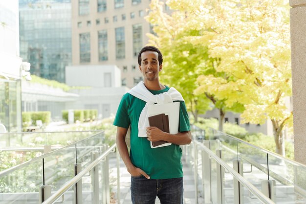 Photo young smiling african american student holding documents notebook wearing stylish green t shirt looking at camera standing outdoors on urban city background successful businessman business concept