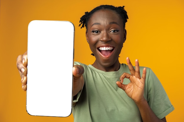 Young smiling african american woman showing smartphone with blank screen against yellow background