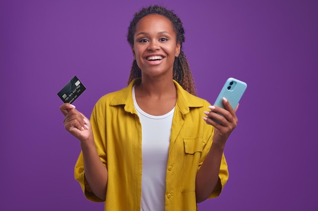 Young smiling african american woman with credit card and phone stands in studio