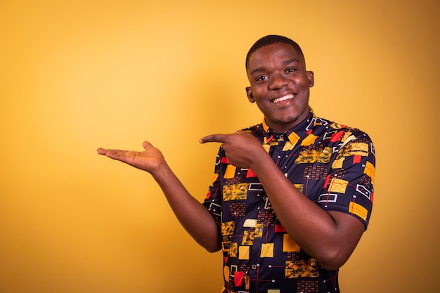 Young smiling African boy isolated on orange background wearing African patterned shirt his finger points to the space on his hand advertising photo with copy space