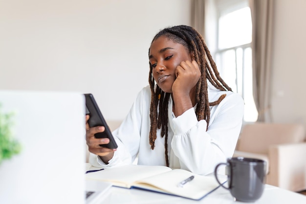 사무실에서 컴퓨터 근처에서 스마트폰을 사용하여 웃고 있는 젊은 아프리카 비즈니스 여성