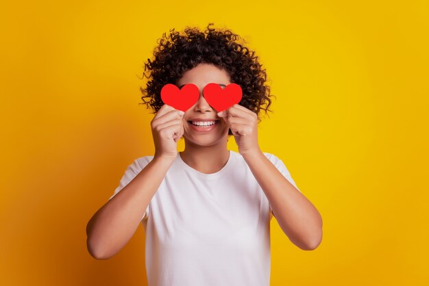 Young smiling african girl hold two little paper heart figures cover eyes wear white t-shirt on yellow wall