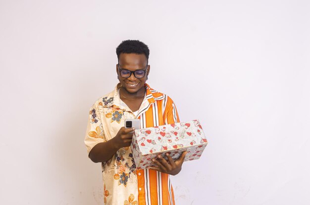 Фото Молодой улыбающийся африканский мужчина держит подарочную коробку и использует свой телефон для покупок в интернете