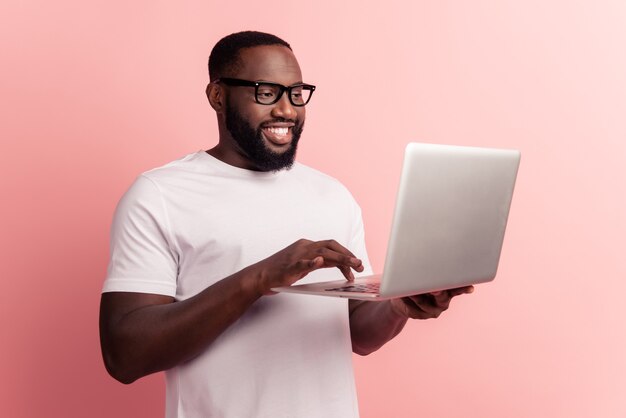 Young smiling african man standing and using laptop computer chatting on pink wall