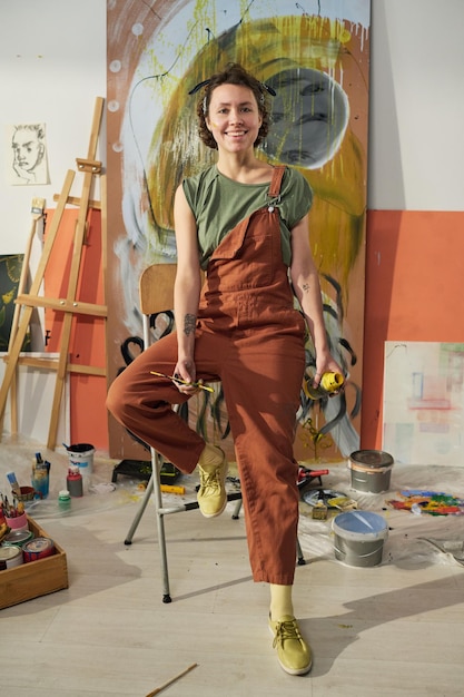 young smiling artist or painter in jumpsuit sitting in the center of studio