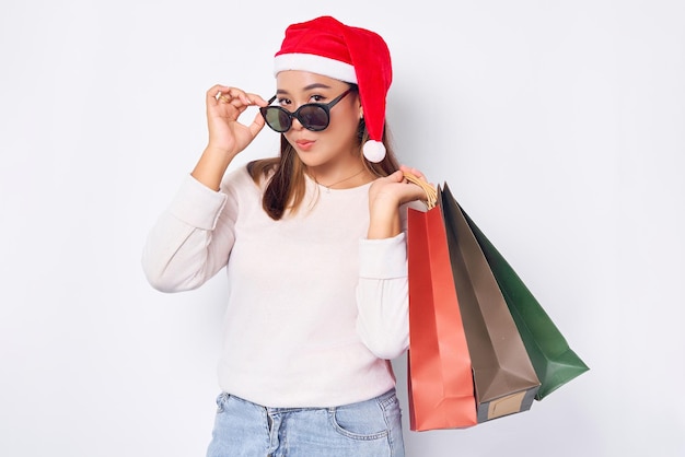 Giovane donna bionda asiatica sorridente degli anni '20 che indossa un cappello di natale che tiene le borse della spesa dopo lo shopping per gli accessori natalizi decollare gli occhiali su sfondo bianco felice anno nuovo natale 2023