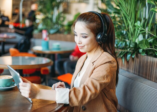 La giovane studentessa asiatica sorridente in cuffie comunica tramite tablet impara la lingua straniera al caffè