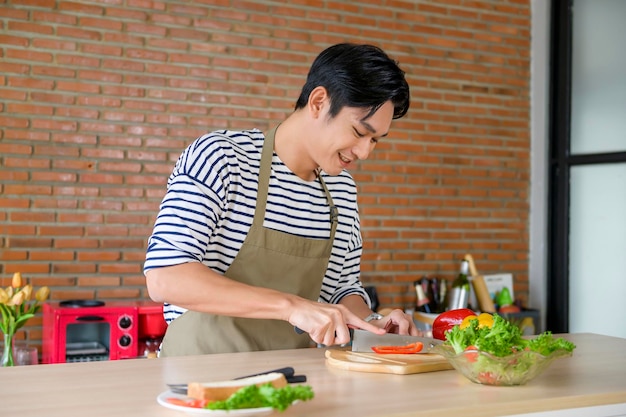 Молодой улыбающийся азиатский мужчина в фартуке на кухне готовит концепцию приготовления пищи