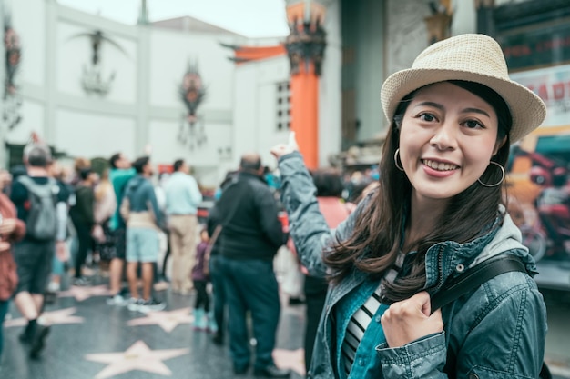 young smiling asian traveler woman in straw hat finger point showing background of famous chinese theatre on hollywood walk of fame. beautiful happy backpacker girl in spring holidays travel usa.