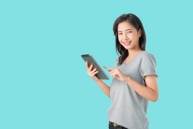 Young smiling Asian woman holding digital tablet with pointing finger on light blue background.