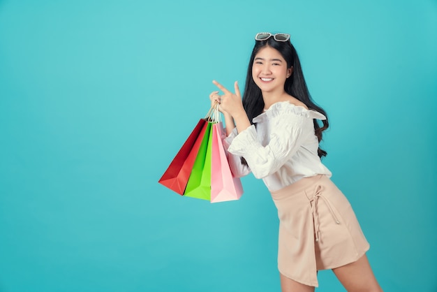 Young smiling asian woman holding multi coloured shopping bags and hands pointing finger