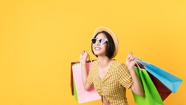 Young smiling asian woman holding multi coloured shopping bags and looking on light yellow wall