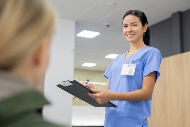 Photo young smiling assistant in blue uniform making notes in registration document while looking at one of patients of dental clinics