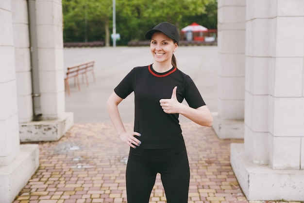 Giovane sorridente atletica bella donna bruna in uniforme nera e berretto facendo esercizi sportivi, riscaldamento prima di correre, mostrando pollice in su nel parco cittadino all'aperto