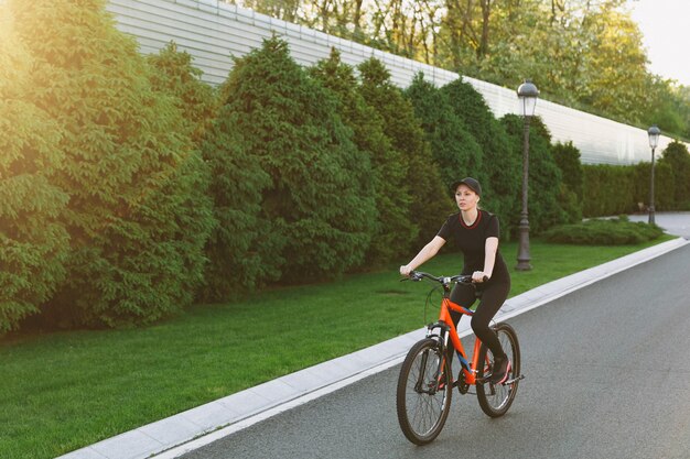 Young smiling athletic brunette strong woman in black uniform, cap riding road on black bicycle with orange elements outdoors on spring or summer sunny day. Fitness, sport, healthy lifestyle concept.