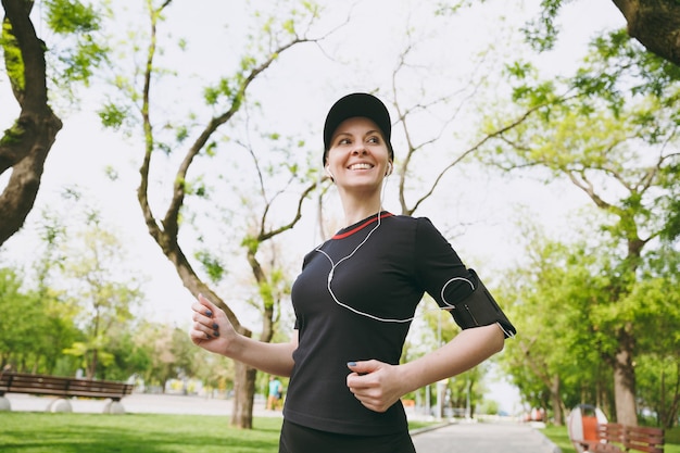 Giovane donna bruna atletica sorridente in uniforme nera e berretto con auricolari che si allenano facendo sport, correndo e ascoltando musica sul sentiero nel parco cittadino all'aperto