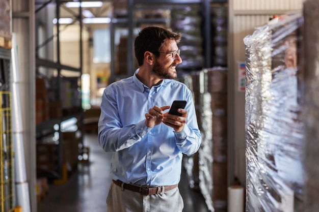 Young smiling attractive bearded businessman standing in warehouse and using smart phone.