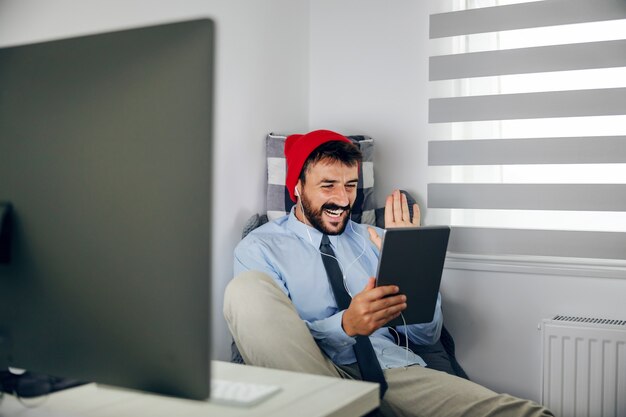 Young smiling attractive bearded businessman with cap on head sitting in his office and having video call over tablet.