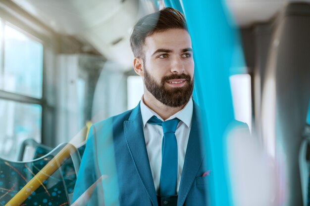 Young smiling attractive businessman in blue suit sitting in public bus and looking away.