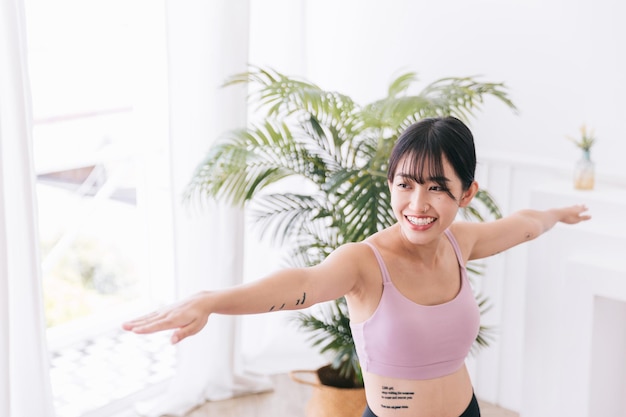 若い笑顔の魅力的なスポーティなアジアの女性がVirabhadrasana2の練習をしてヨガを練習し、戦士2のポーズで屋内で運動し、スポーツウェアを身に着けているハーフレングスのボディショット