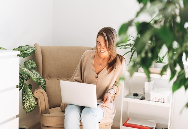 Young smiling attractive woman in cozy beige cardigan working at laptop sitting in chair at the home with green house plants