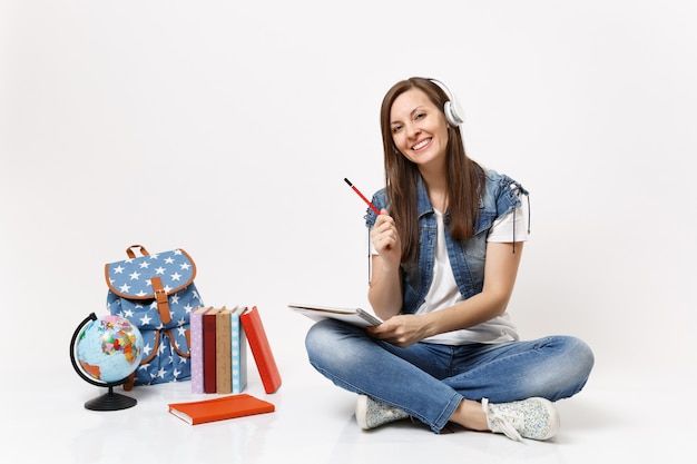 ヘッドフォンで若い笑顔の魅力的な女性の学生は、ノートブック、地球の近くの鉛筆、バックパック、孤立した教科書を保持している音楽を聴きます