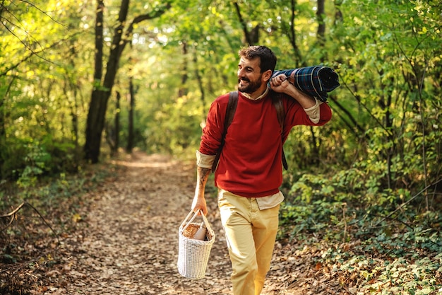 Young smiling bearded man walking in nature, having backpack on backs and holding picnic equipment.