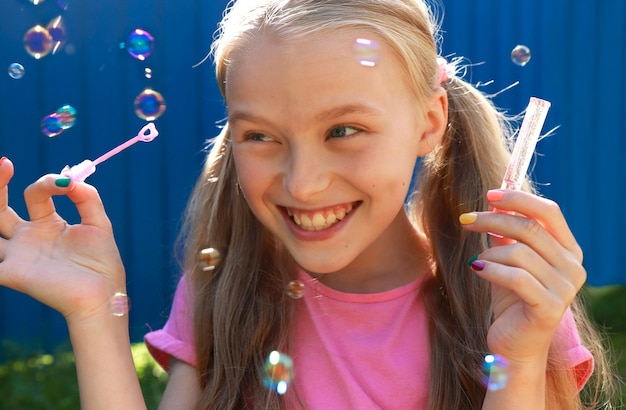 A young smiling beautiful blonde girl inflates soap bubbles in the summer on a blue background.