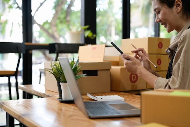 Young smiling beautiful owner asian woman freelance sme business online shopping working on laptop computer with parcel box on desk at home SME business online and delivery concept