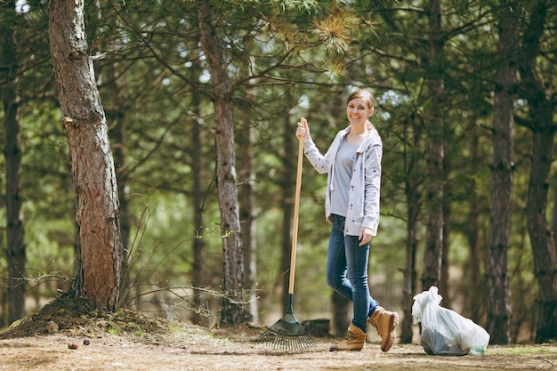 Young smiling beautiful woman cleaning and using rake for garbage collection near trash bags in park or forest. Problem of environmental pollution