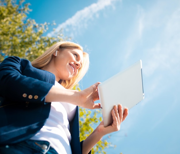 Young smiling beautiful woman in park with tablet gadget and wireless headphones enjoying green energy concept remote work