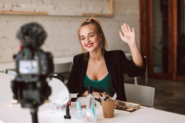 Young smiling beauty blogger happily waving on camera while recording new make up video for her vlog