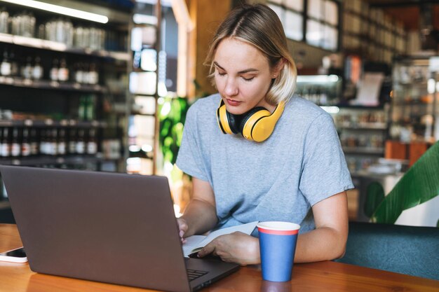 オンラインコースを勉強しているカフェの女の子の学生のテーブルでノートに取り組んでいる黄色のヘッドフォンで若い笑顔のブロンドの女性フリーランサー