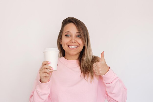 Young smiling blonde woman holding white eco paper cup with tea or coffee showing thumb up gesture