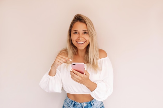 Young smiling blonde woman holds mobile phone in her hand and points at the screen with her finger