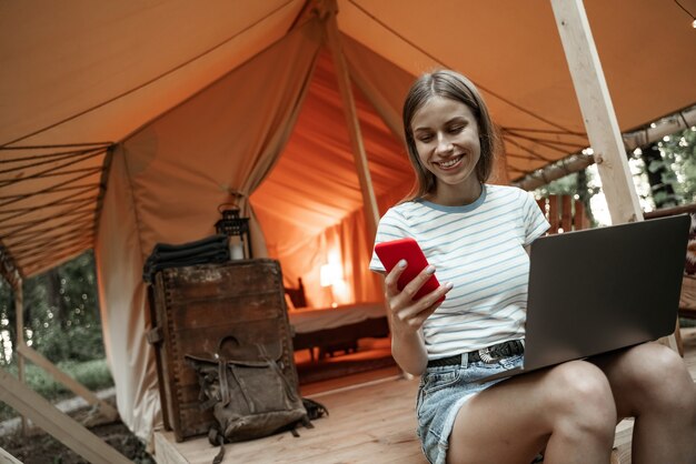 Young smiling blonde woman sitting on glamping ground using laptop and messaging on smartphone. Camping lifestyle. Low-budget travel. Remote work concept. Modern internet communication technology.