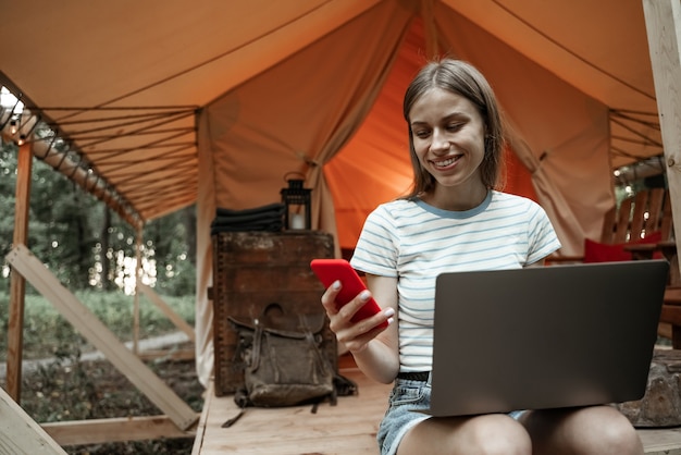 Young smiling blonde woman sitting on glamping ground using laptop and talking on smartphone. Camping lifestyle. Low-budget travel. Remote work concept. Modern internet communication technology.