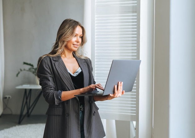 Young smiling blonde woman with long hair in stylish grey suit working at laptop in the bright modern office