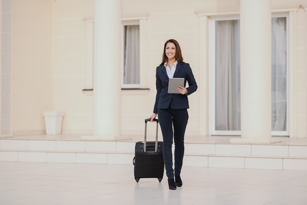 Young smiling brunette dressed smart casual walking, holding luggage and tablet. Business trip concept.