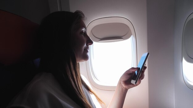 ゆるい髪の若い笑顔のブルネットは飛行機の近くのビューで明るい舷窓に対して電話サーフィンインターネットを見て