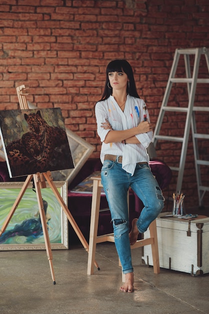 彼女のスタジオで笑顔の若いブルネットの女性アーティストがブラシを持っています。