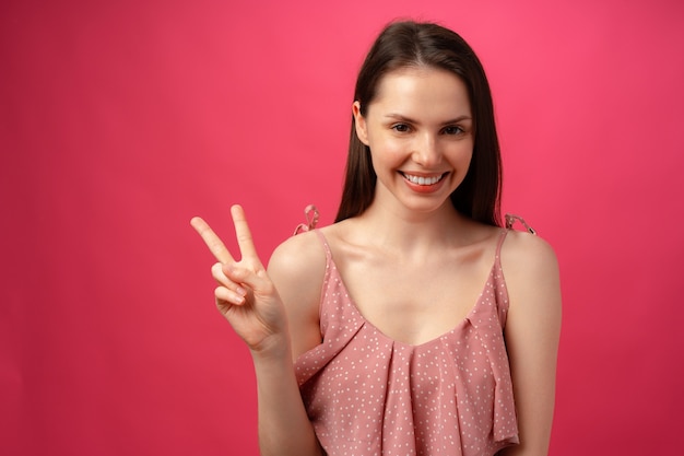 Молодая улыбающаяся брюнетка женщина показывает знак победы на розовом фоне