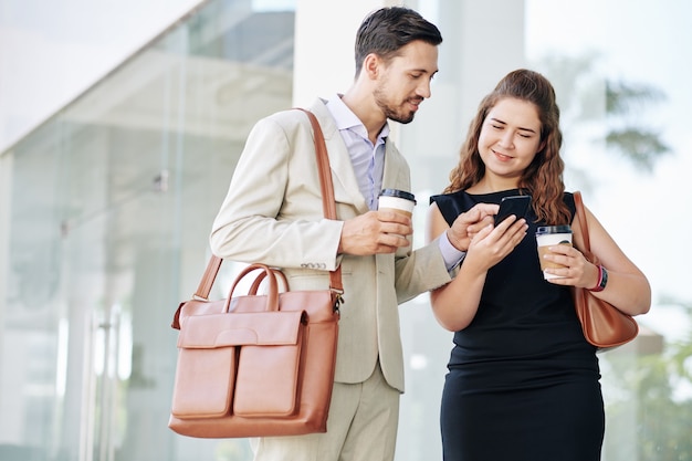 Young smiling business people with take out coffee discussing funny video or meme on smartphone screen when standing in the street