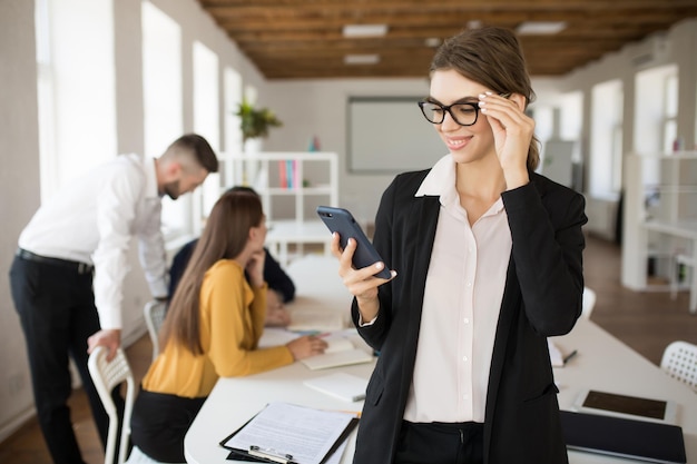 Young smiling business woman in eyeglasses and shirt happily using cellphone while spending time in office with colleagues on background