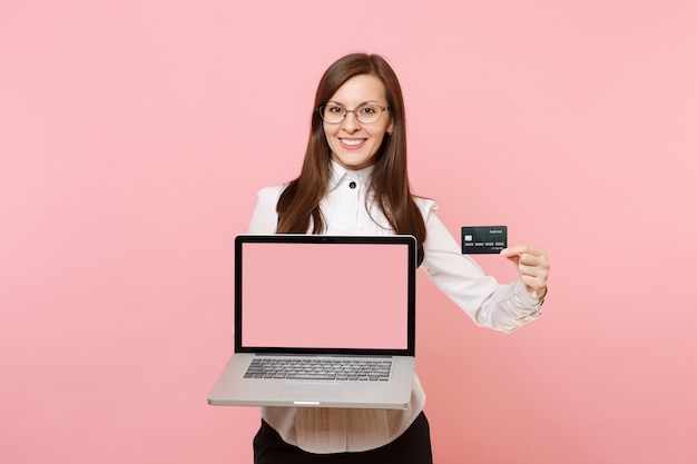 Young smiling business woman in glasses holding credit card, laptop pc computer with blank empty screen isolated on pink background. Lady boss. Achievement career wealth. Copy space for advertisement.